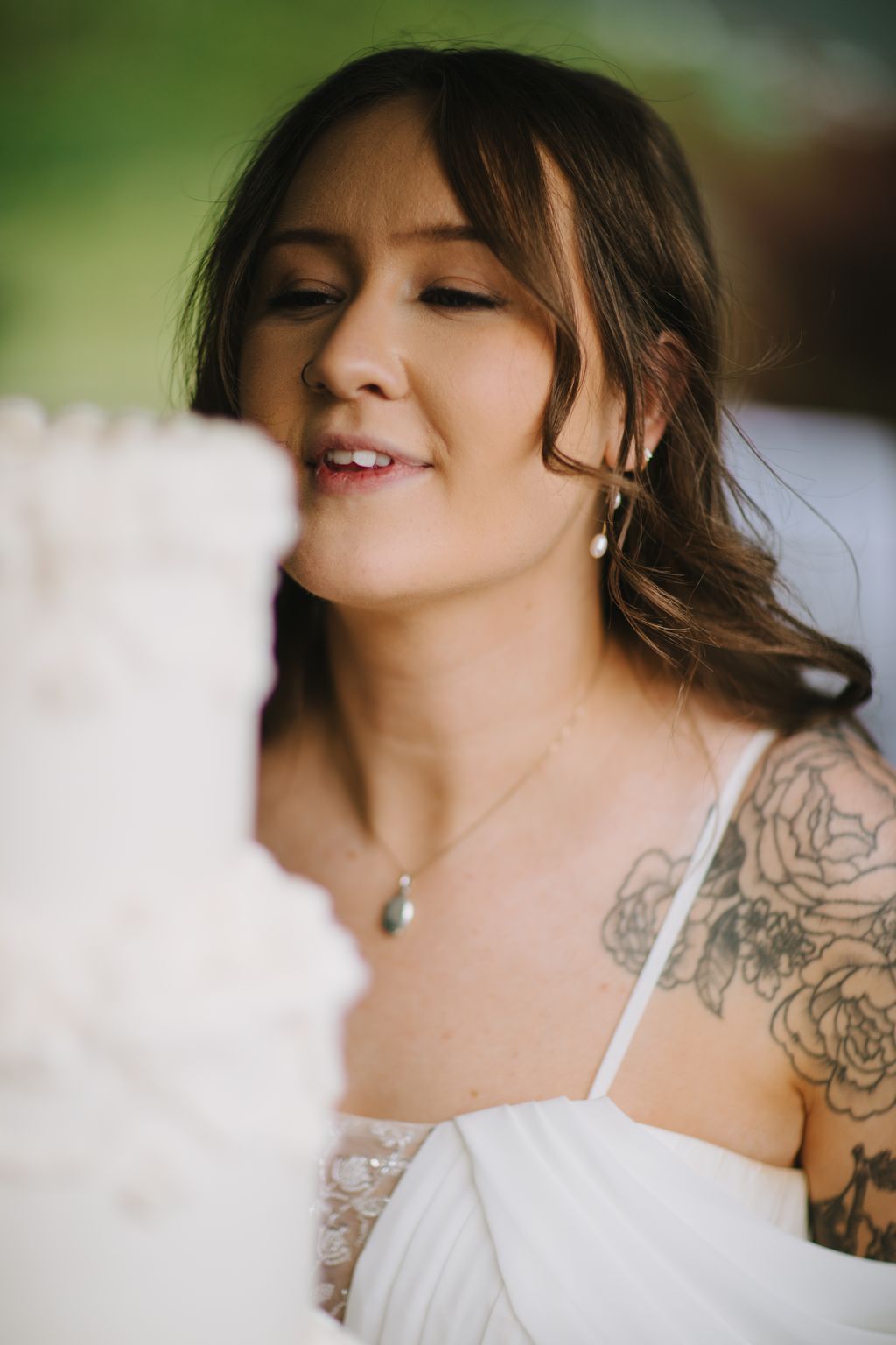 Bride with dark hair and bangs smiling with eyes closed, wearing pearl earrings and white off-shoulder dress against soft green background