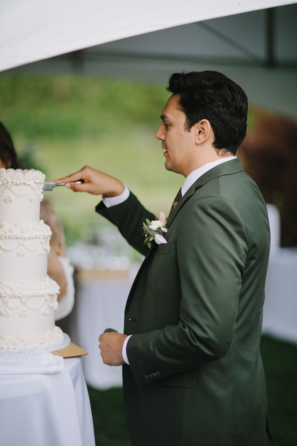 Groom in olive suit with boutonniere gestures while speaking outdoors under tent on wedding day