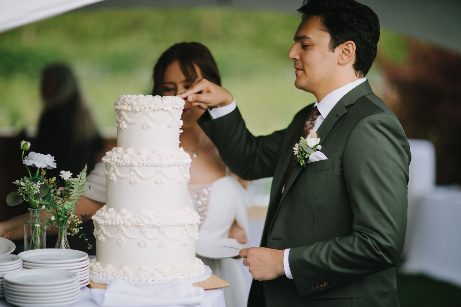 Bride and groom cut into their white tiered wedding cake outdoors with soft green background