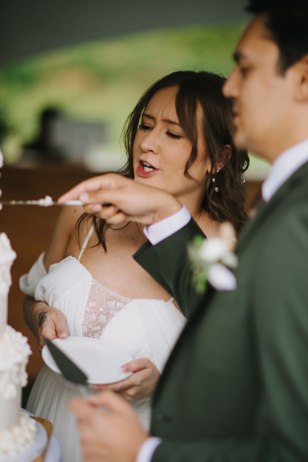 Bride in white lace dress smiling during outdoor ceremony with blurred green background and groom visible beside her