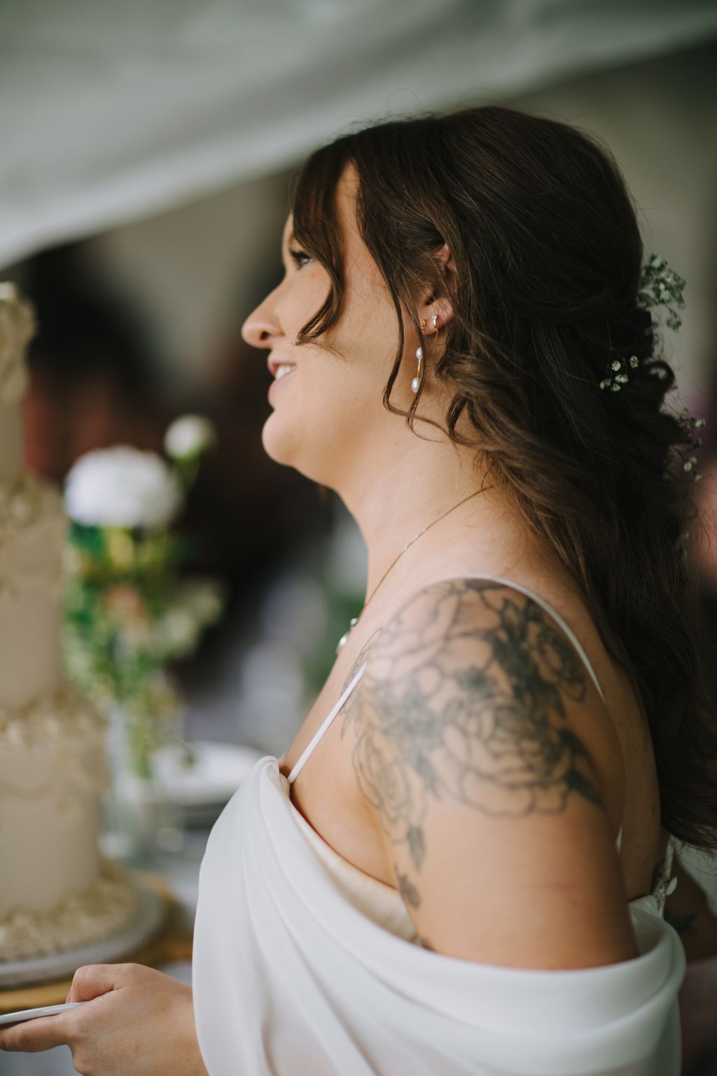 Bride in profile wearing white dress with shoulder tattoo visible, soft natural light, gentle smile, blurred green background