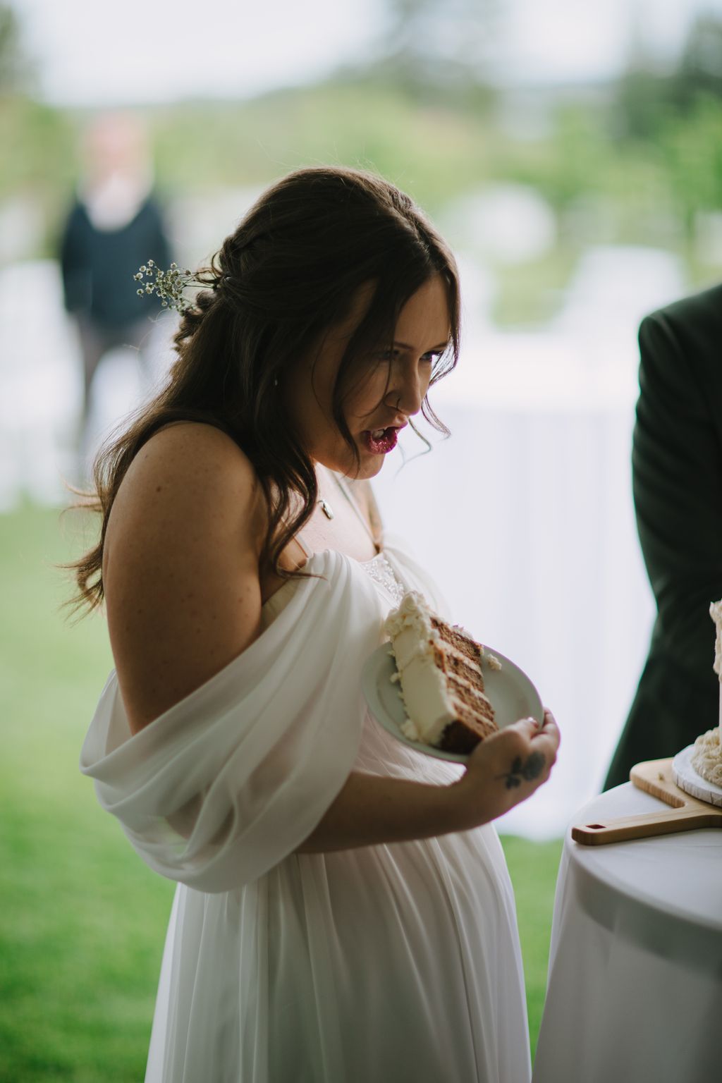 Bride in white dress holding layered cake slice, smiling in profile with blurred green outdoor background