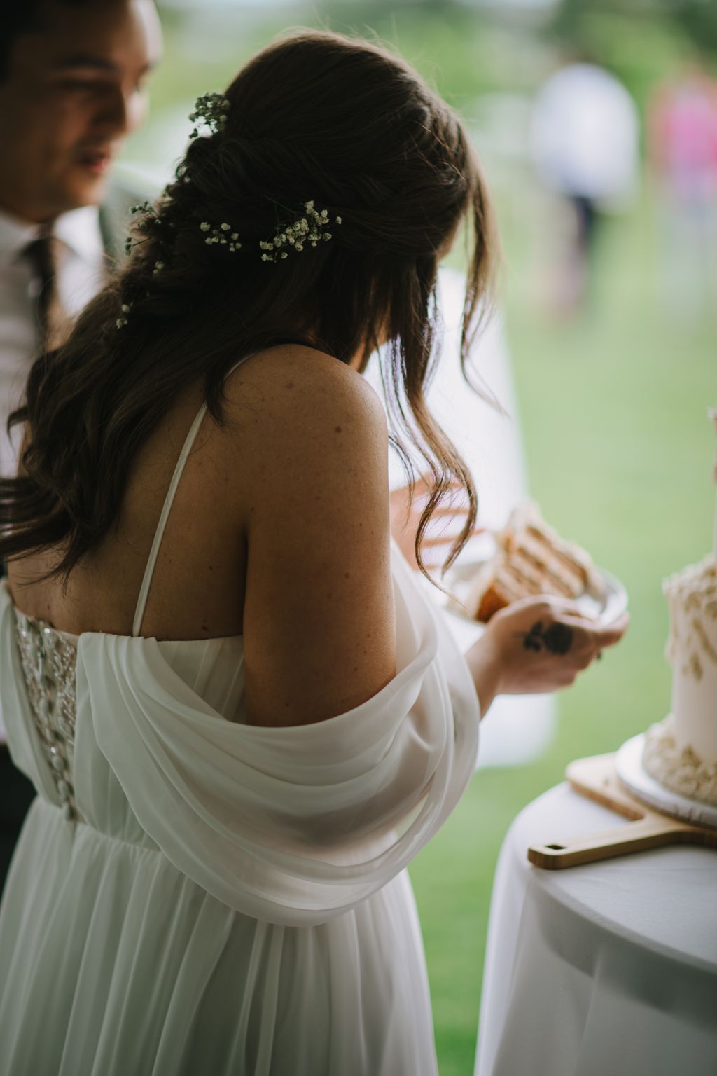 Bride in white dress viewed from behind, dark hair adorned with baby''s breath flowers, soft outdoor garden setting with blurred green background