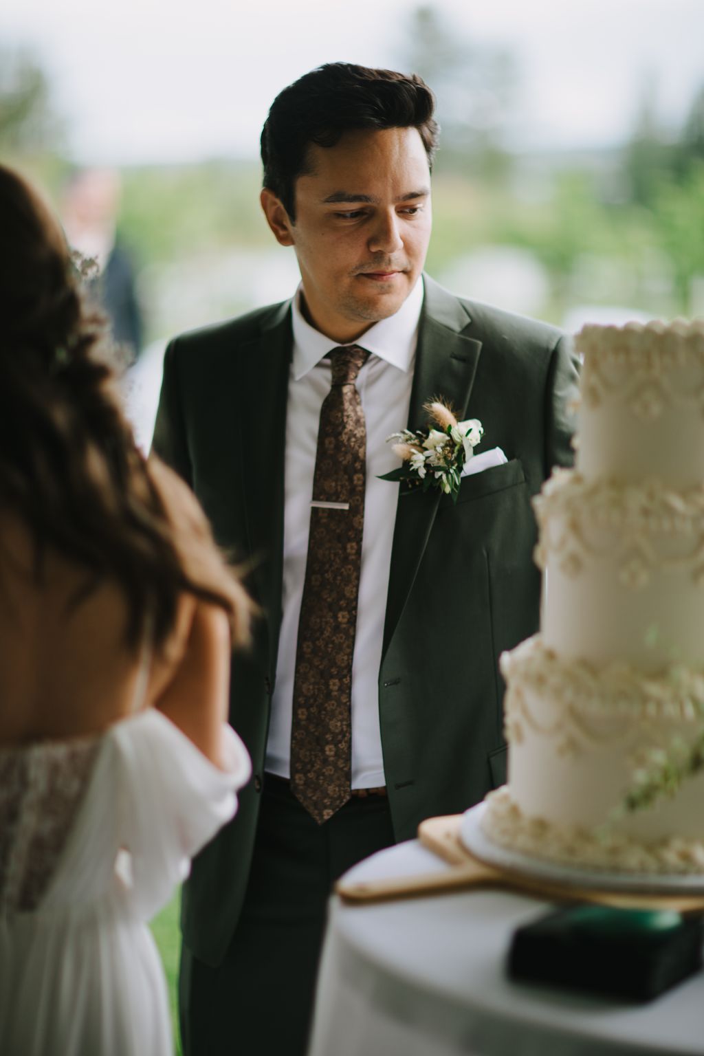 Groom in charcoal suit and brown tie with boutonnière gazes downward during ceremony outdoors