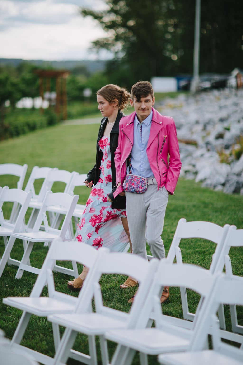 Couple walking through outdoor ceremony seating, woman in floral dress and black jacket, man in bright pink blazer and patterned crossbody bag, lush green la…