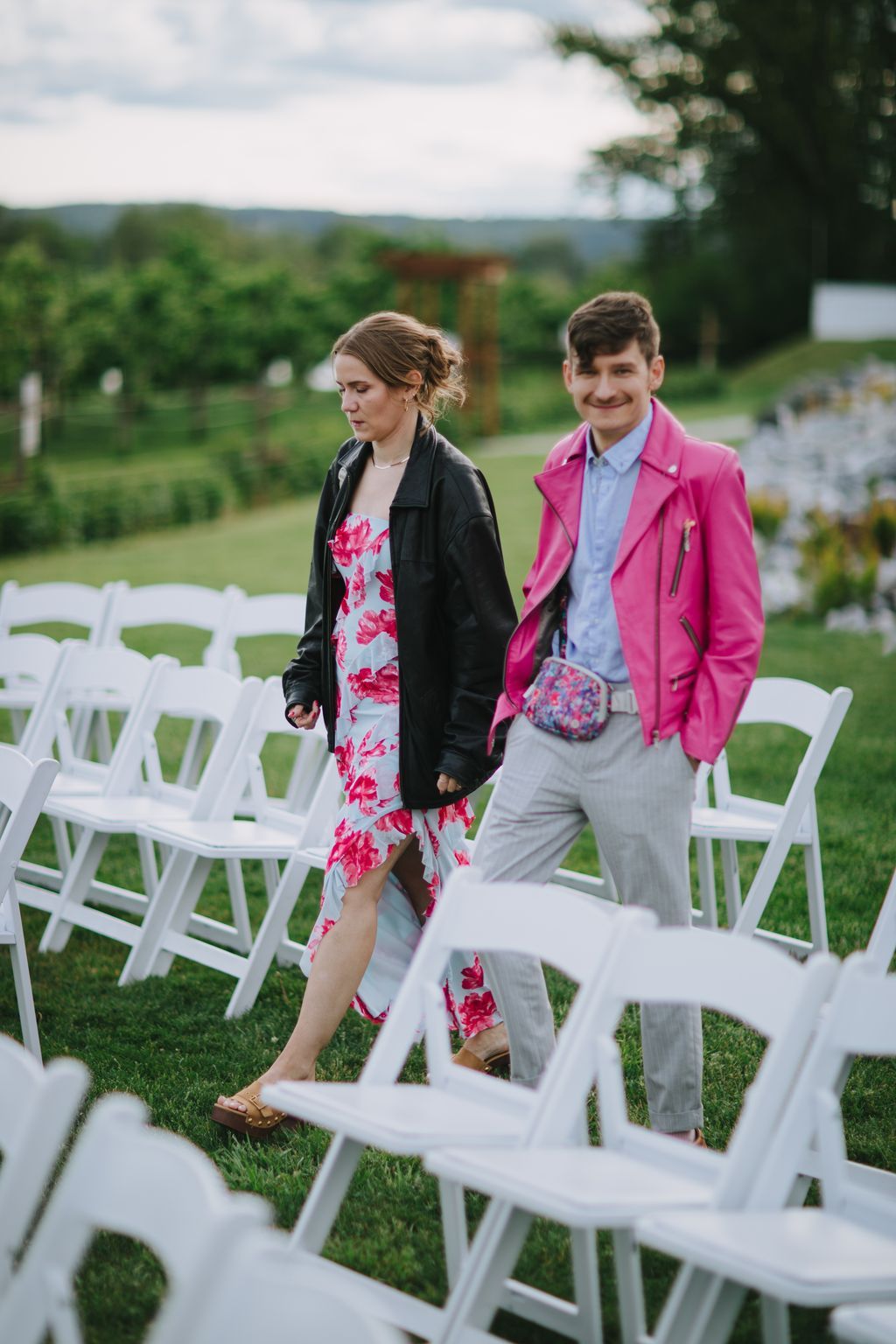 Couple walks through outdoor garden ceremony space with white chairs, bride in floral dress and black jacket, groom in pink blazer and light blue shirt