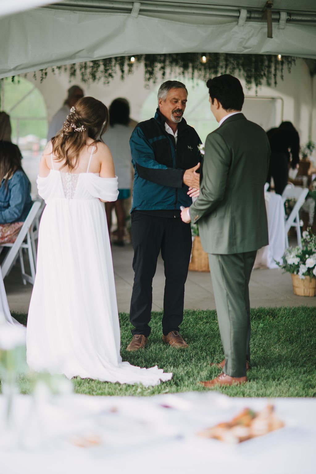 Father of the bride speaks with groom under tent at outdoor wedding reception with hanging greenery