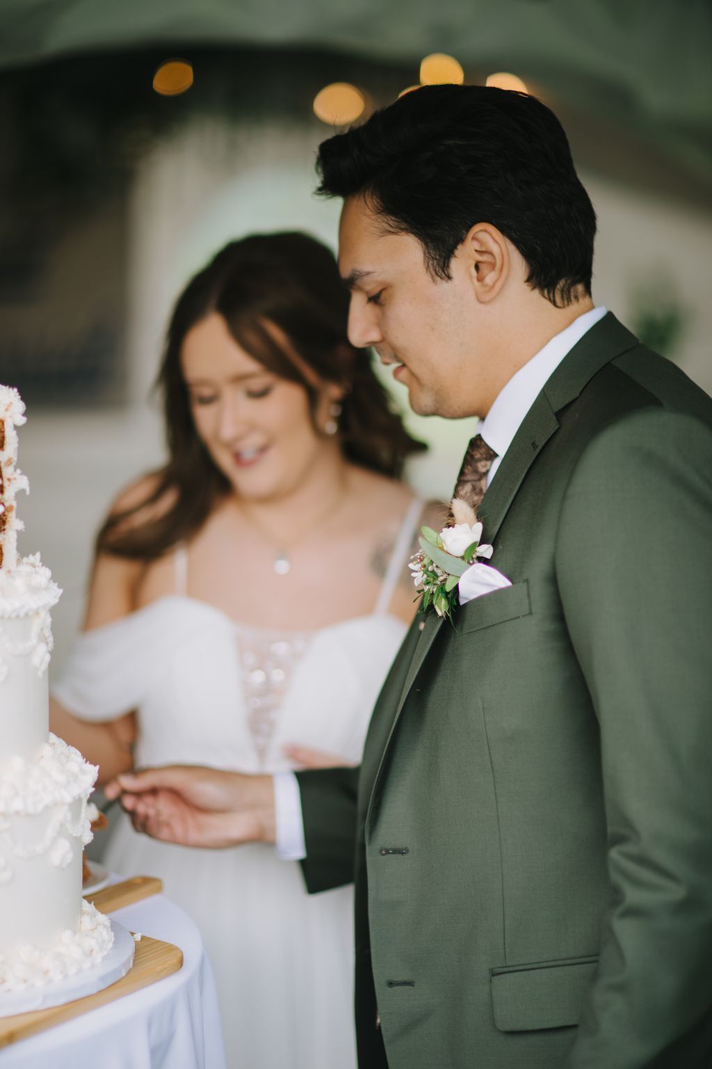 Groom in sage green suit with boutonniere gazes down at bride in white dress during intimate evening ceremony with golden bokeh lights behind them