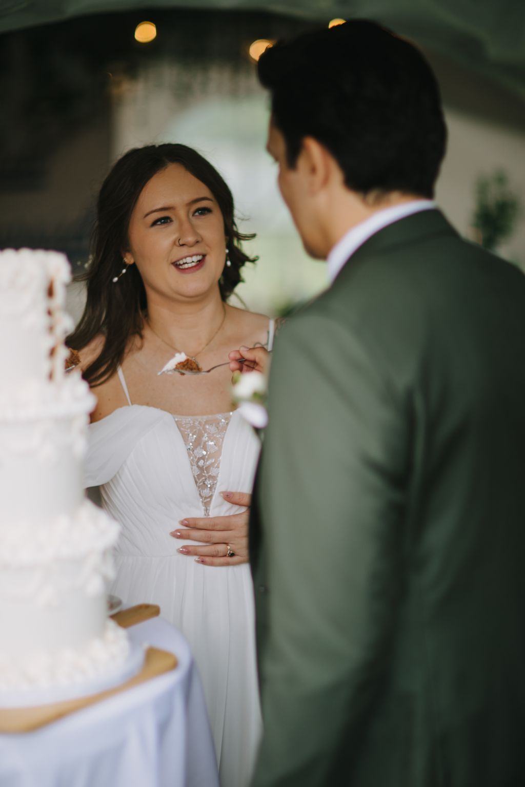 Bride in white beaded gown smiles at groom in sage suit during evening reception with warm bokeh lights behind them