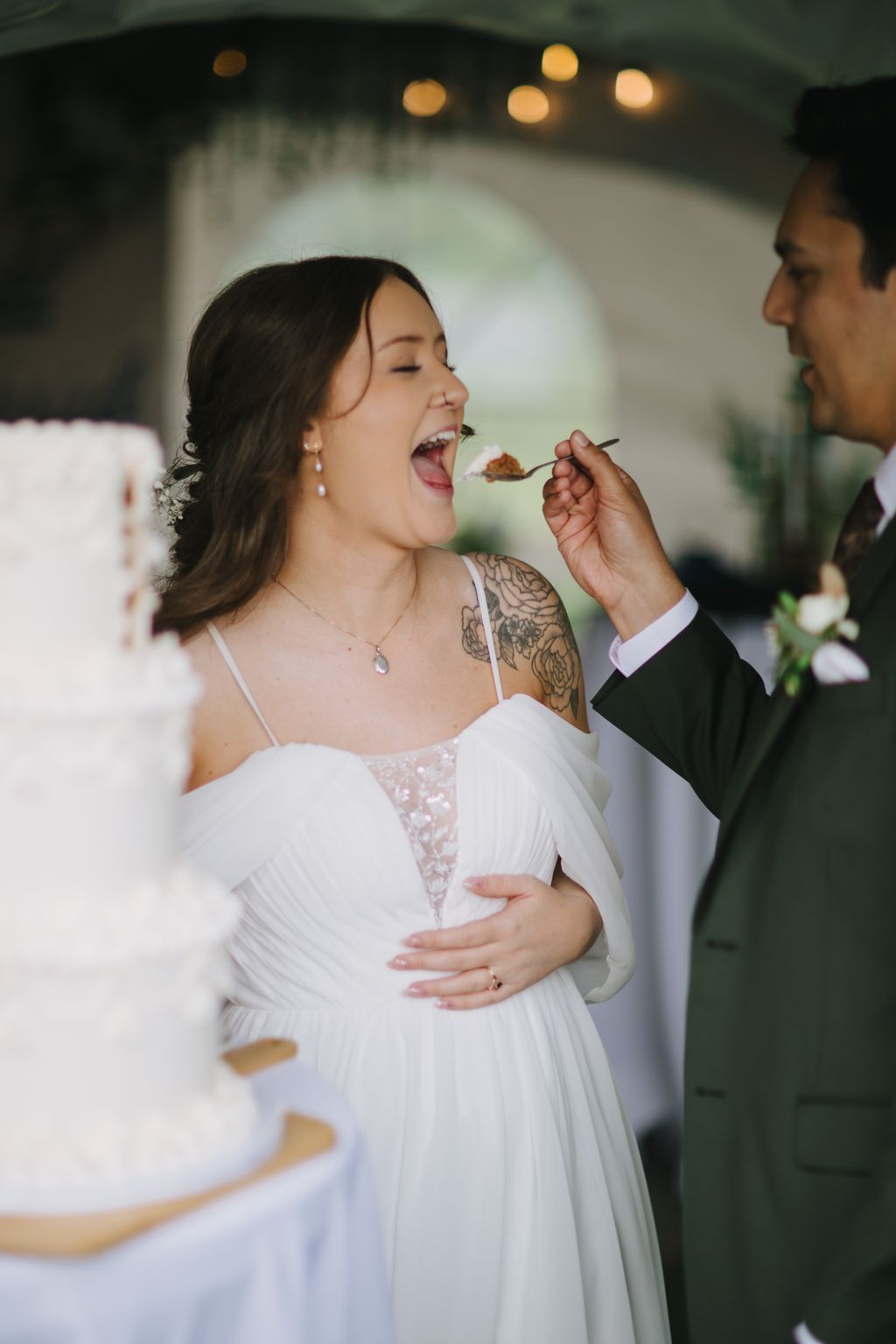 Bride in white dress laughs while eating cake during reception with warm ambient lighting