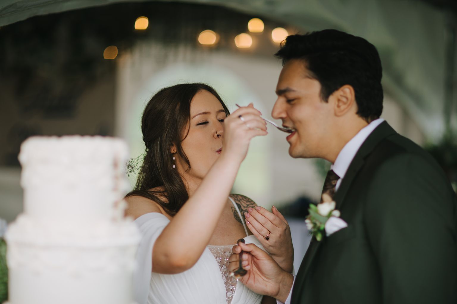 Bride feeds groom cake during reception with warm bokeh lights glowing softly in background