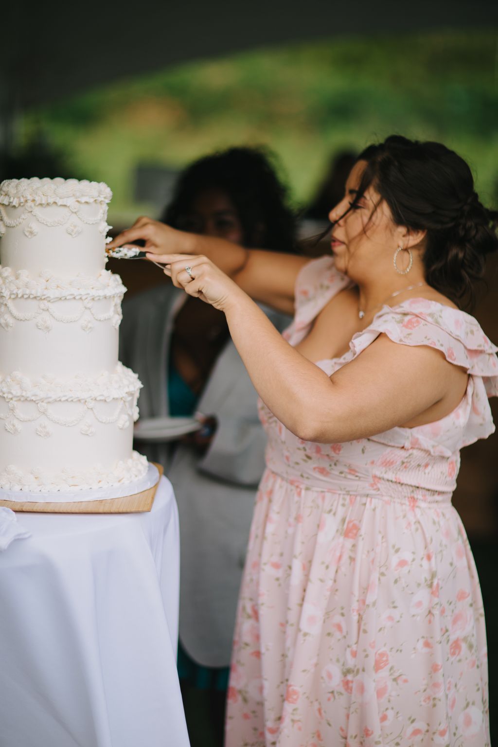 Woman in pink floral dress cuts white wedding cake outdoors with bokeh green background