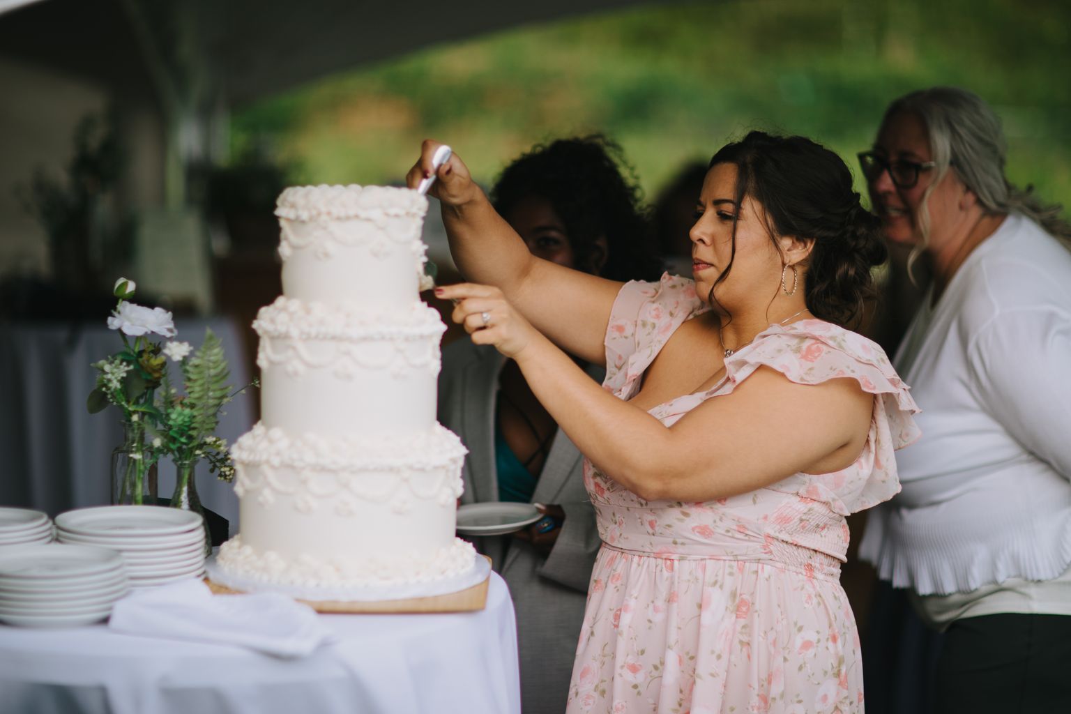 Bride in pink floral dress carefully decorates white wedding cake outdoors with green foliage backdrop