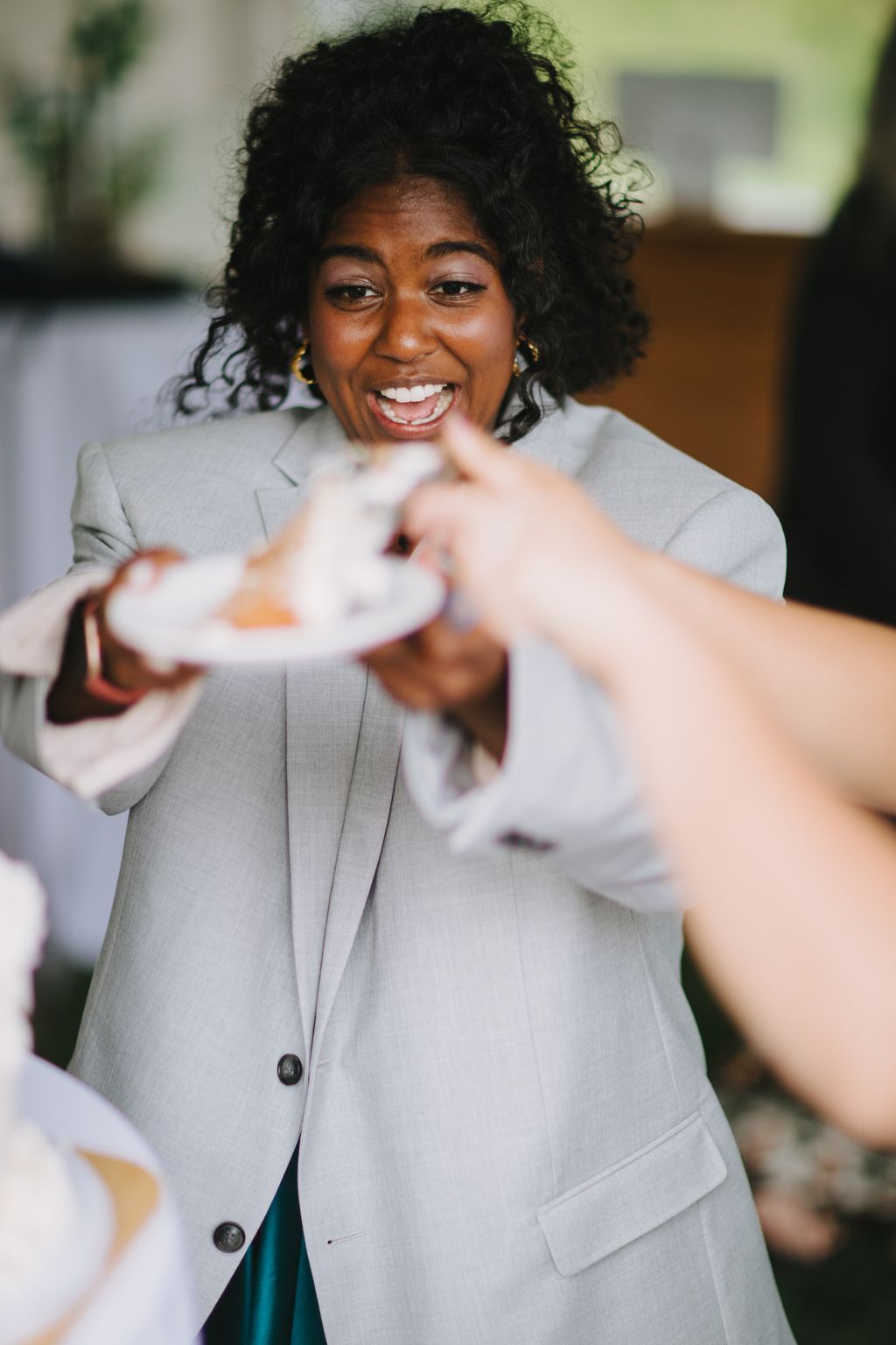 Woman in light gray blazer smiles warmly while holding out her hand, with soft natural lighting and blurred background