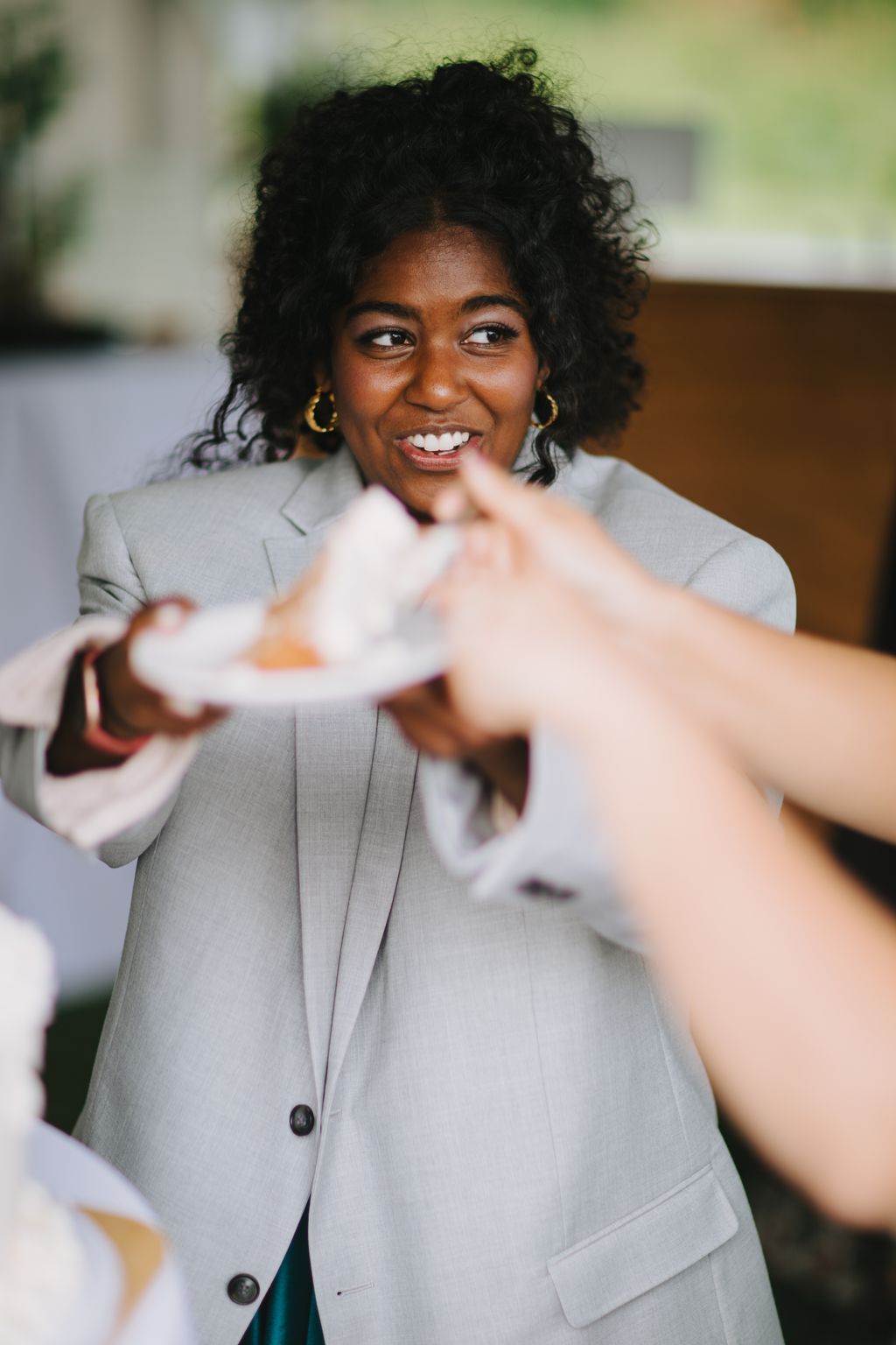 Smiling woman in light gray blazer and gold hoop earrings, engaged in conversation with someone out of frame
