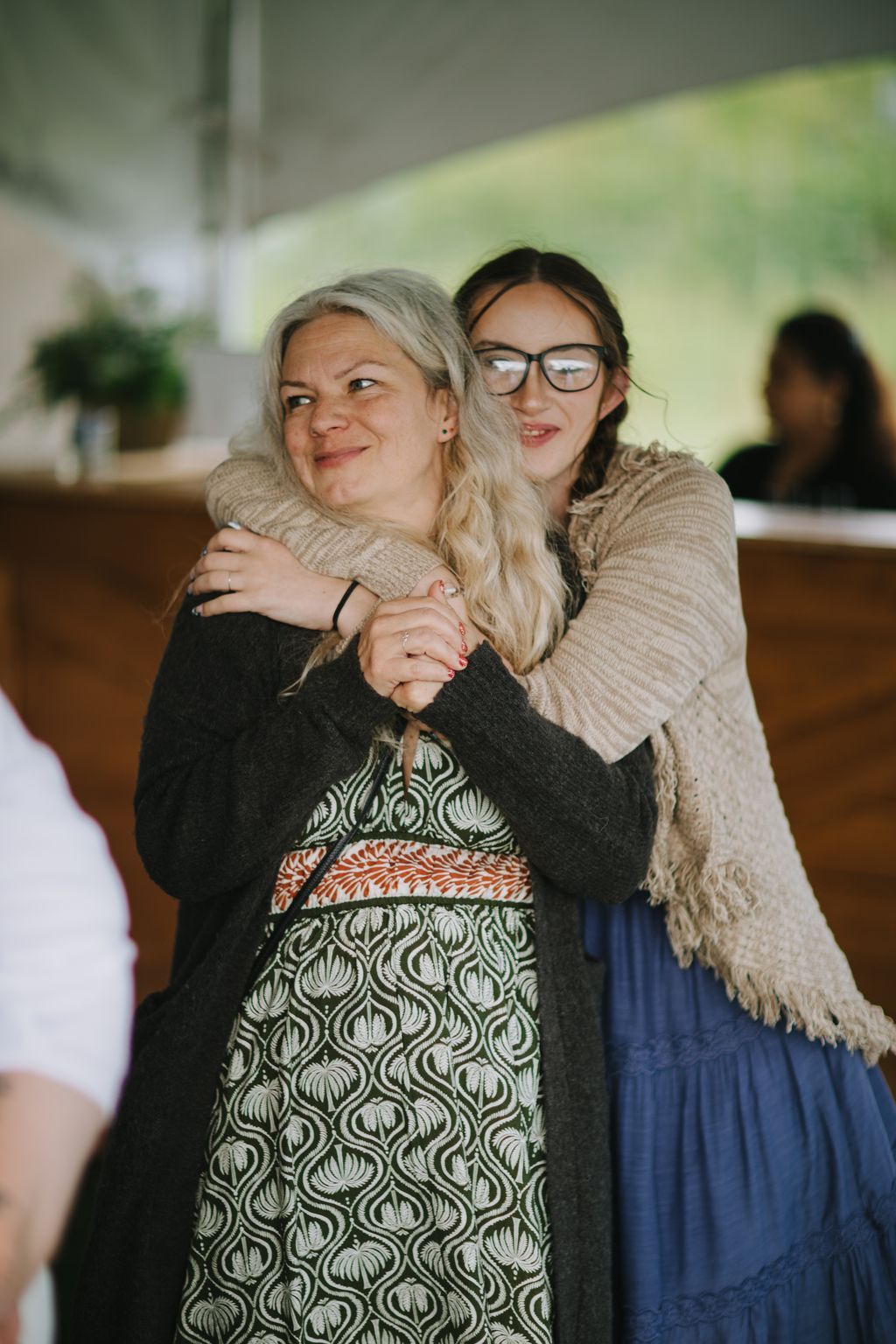 Two women embrace warmly indoors, smiling at the camera in natural light with a soft, blurred green background