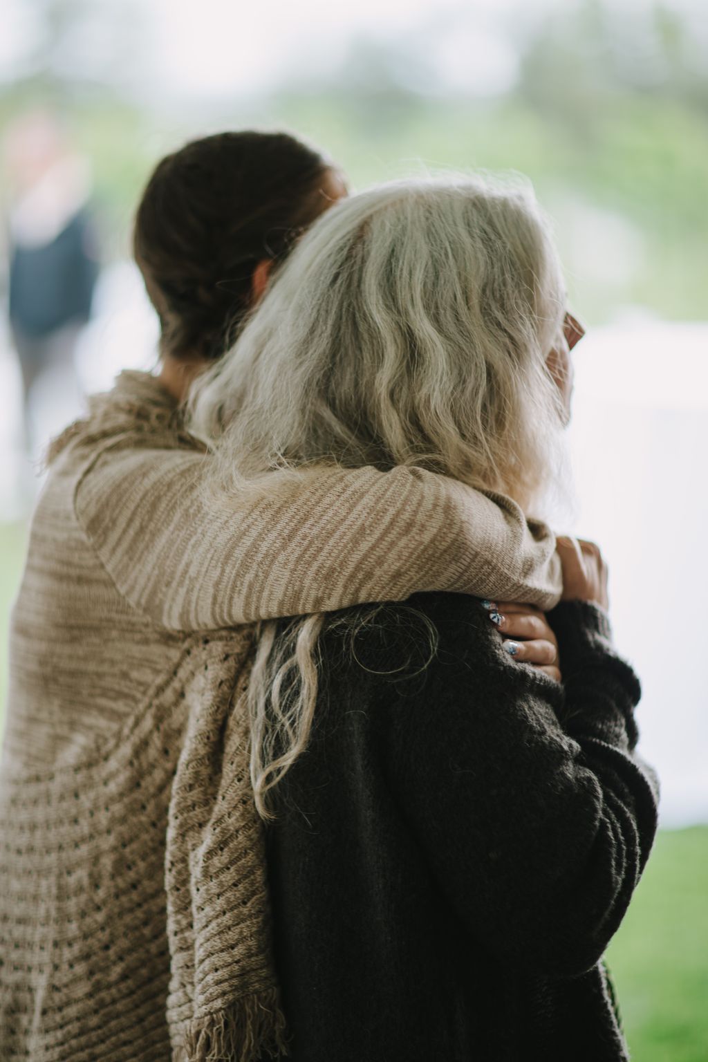 Bride with gray hair embraces groom from behind in soft outdoor light, showing intimate moment and wedding rings