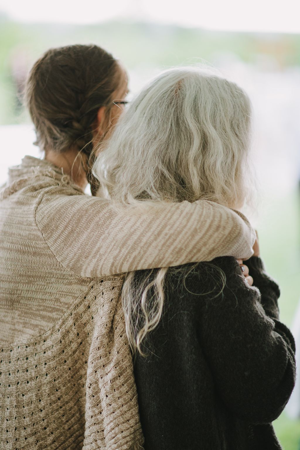 Two people embrace from behind with soft-focus nature background, wearing neutral knit sweaters in warm natural light