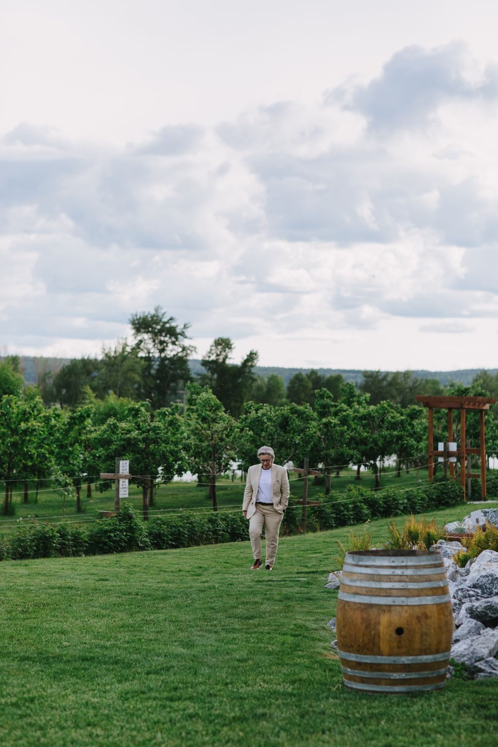 Groom in beige suit walking through vineyard grass with wooden barrel and grapevines under cloudy sky