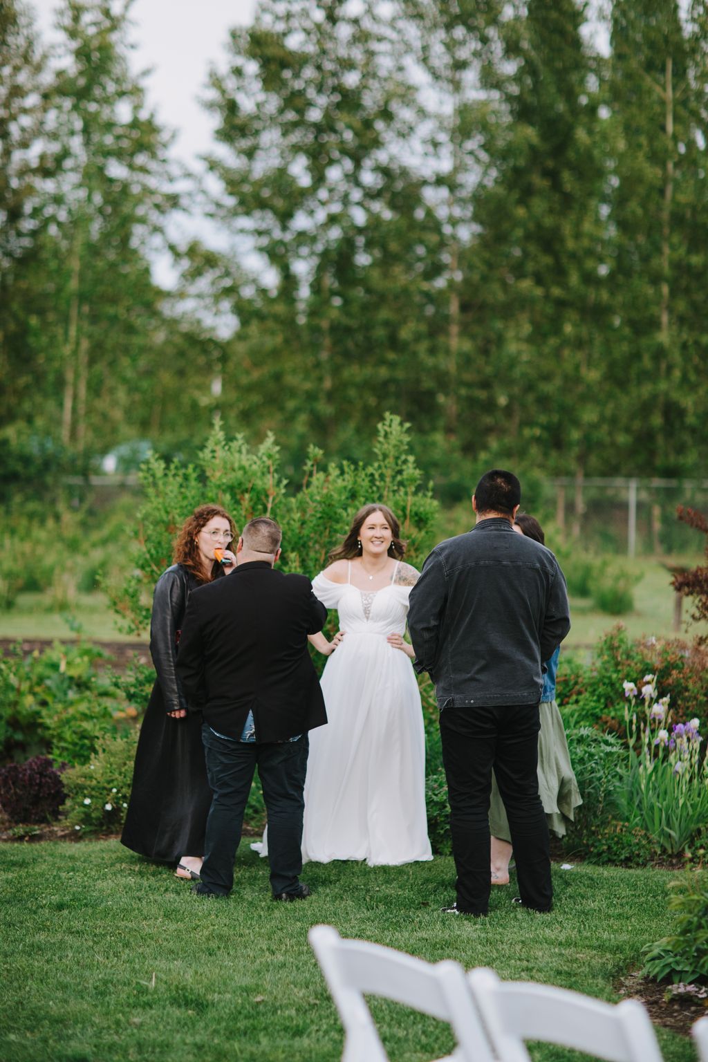 Bride in white dress smiles among guests on grass with blooming trees and greenery in soft daylight