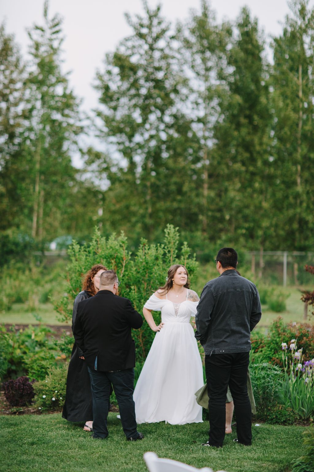 Bride in white off-shoulder gown stands with two guests on grass with blooming trees and hedges behind her