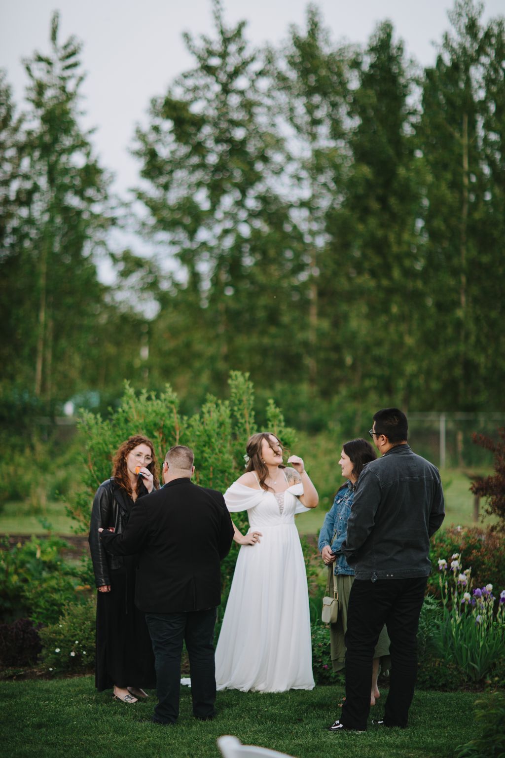 Bride in white off-shoulder gown laughs with guests under blooming trees in a lush garden setting