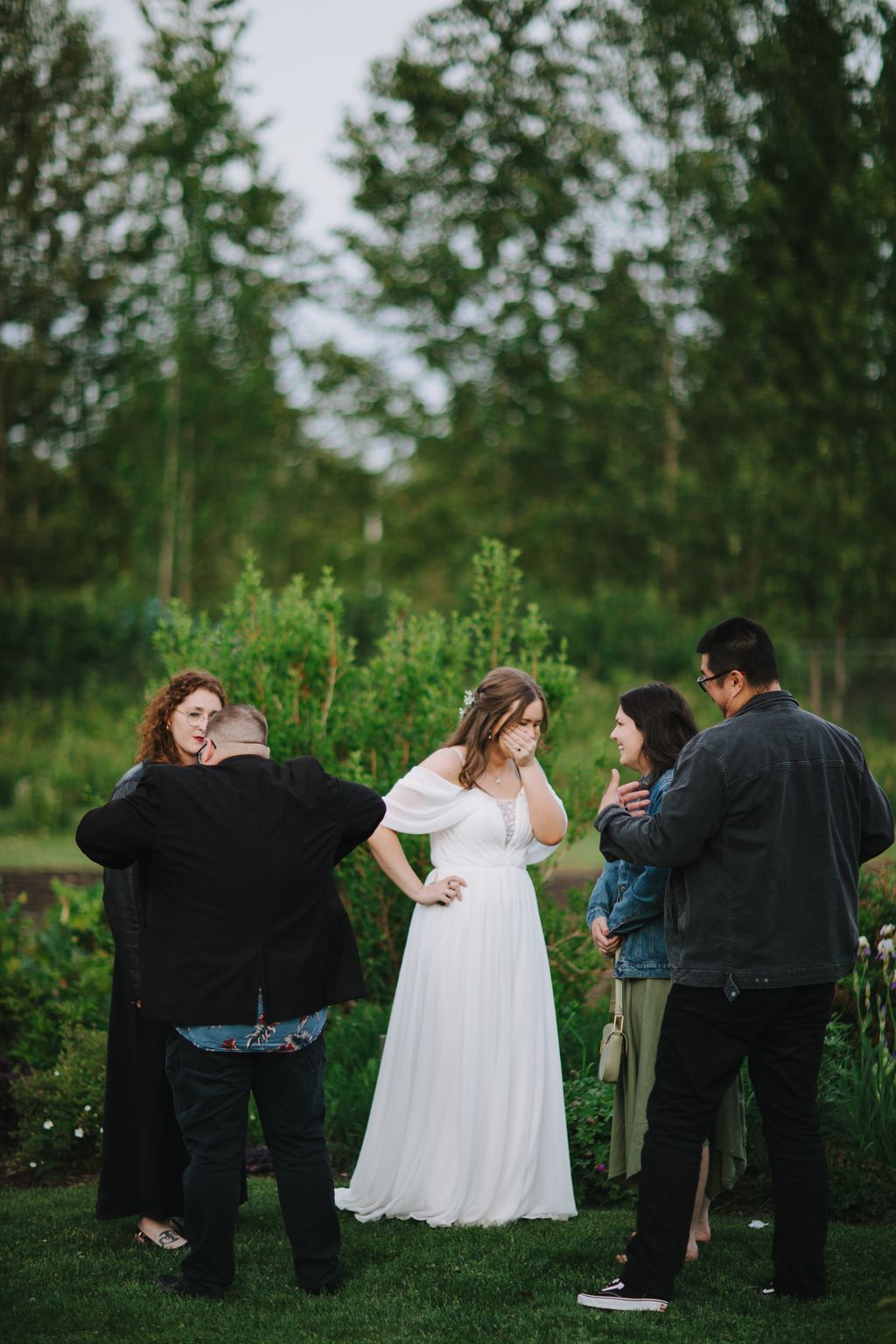 Bride in white off-shoulder gown laughs with guests outdoors under blooming trees on green lawn