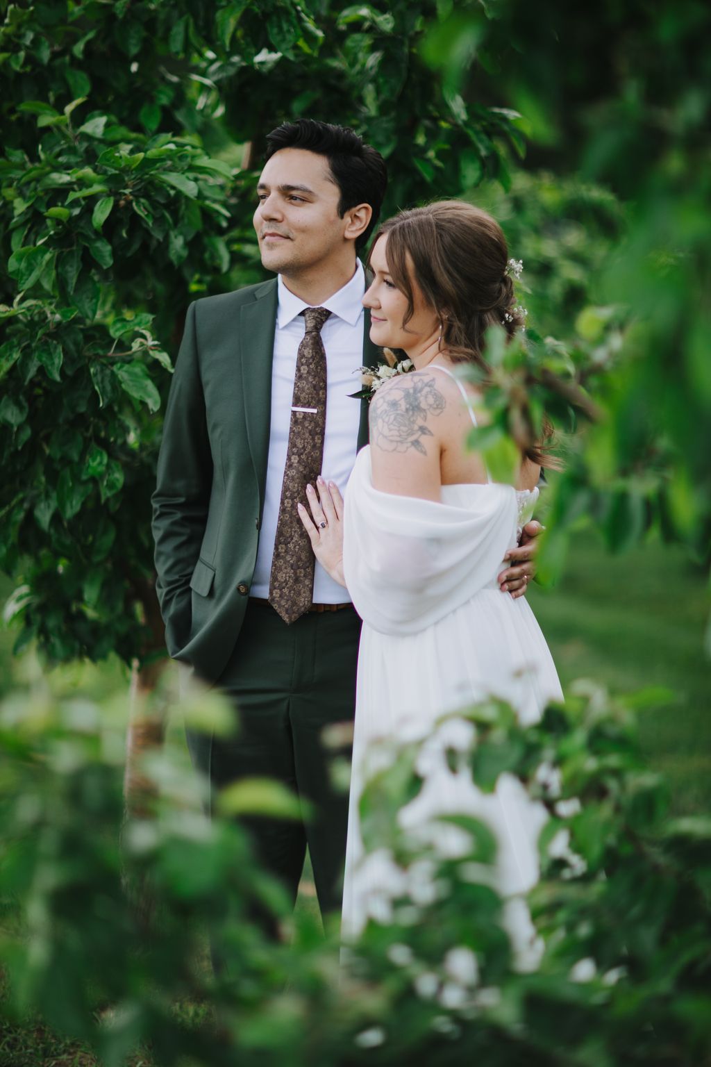 Groom in sage green suit and patterned tie stands with bride in white off-shoulder dress surrounded by lush green foliage and white flowers