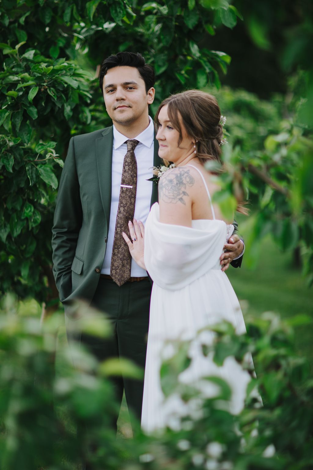 Bride in white off-shoulder gown and groom in sage suit embrace among lush green foliage in natural light
