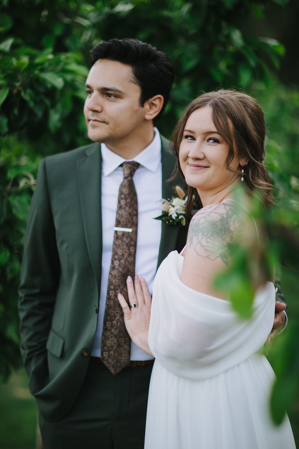 Bride in white dress and groom in sage green suit stand together against blurred green foliage, both gazing thoughtfully away