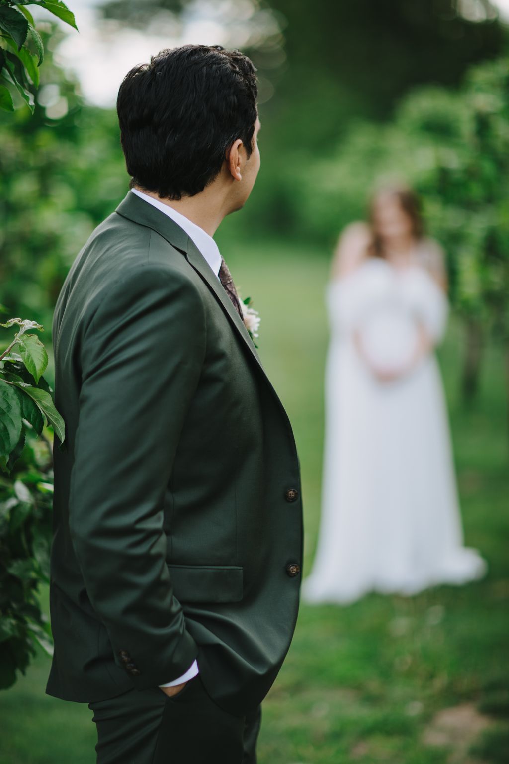 Groom in sage green suit gazes toward bride in white dress during outdoor ceremony, soft natural light