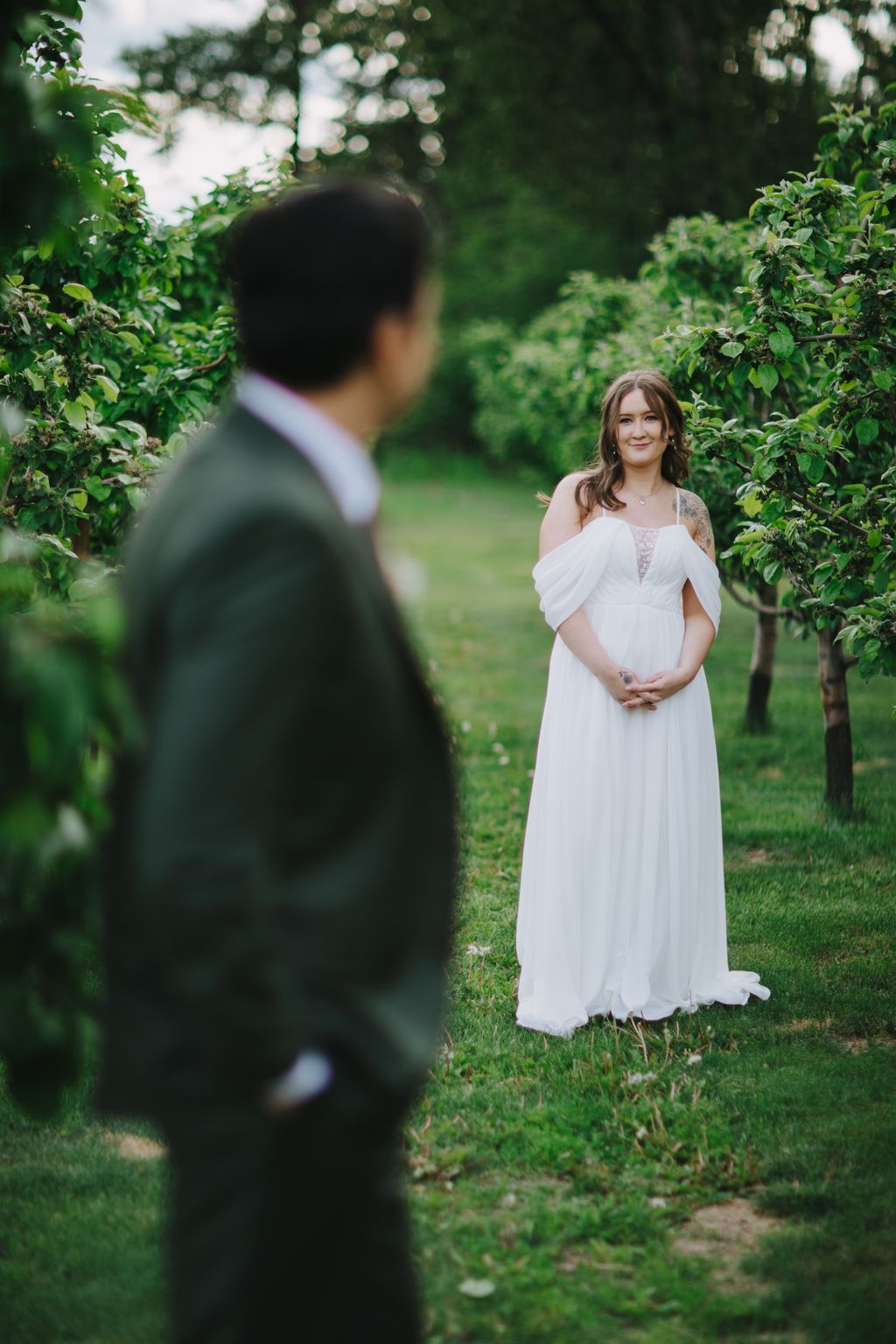 Bride in white off-shoulder gown smiles at groom in dark suit across green lawn under dappled sunlight