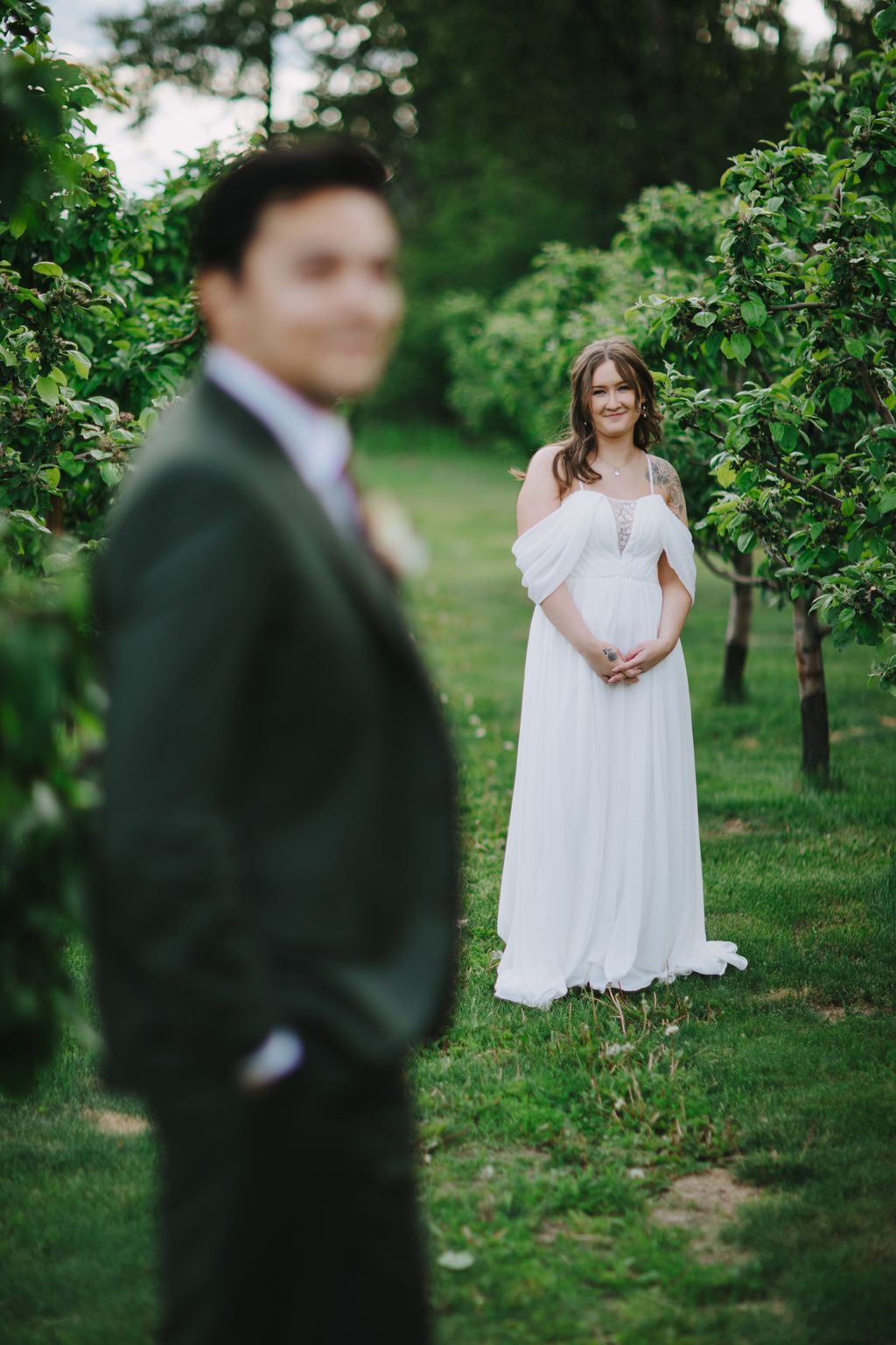 Bride in white off-shoulder gown smiles at camera while groom in dark suit stands blurred in foreground, lush green garden backdrop