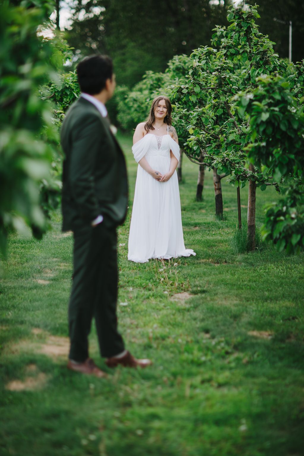 Bride in white cold-shoulder gown smiles at groom in dark suit among lush green orchard trees
