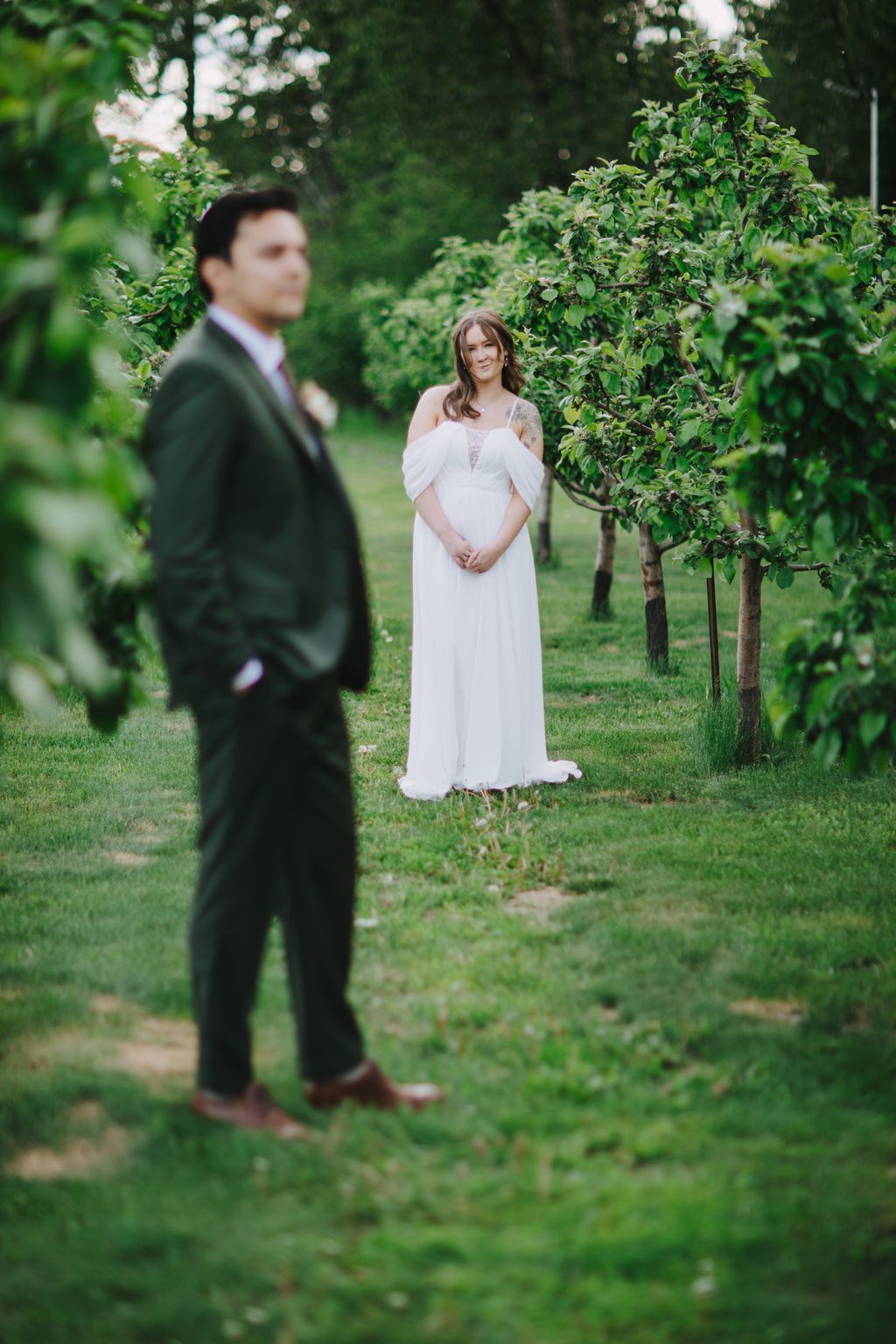 Bride in flowing white off-shoulder gown stands in lush green garden as groom in dark suit approaches from foreground