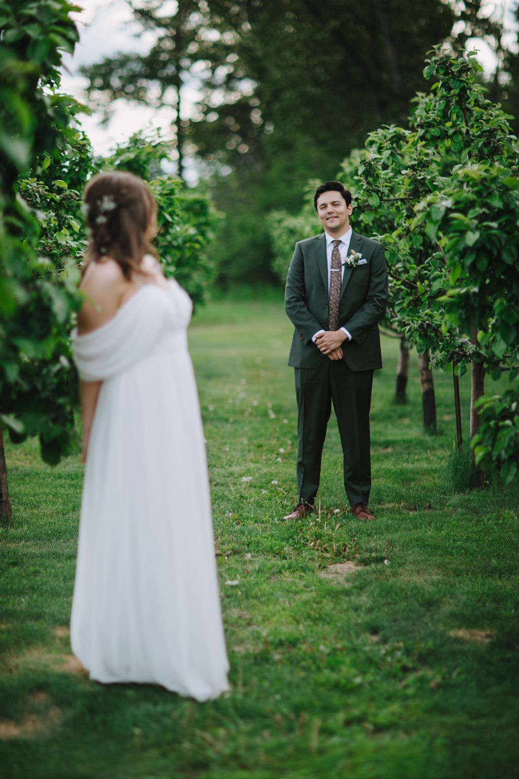 Groom in olive suit smiles while facing bride in white dress in lush green garden setting with soft bokeh foliage background