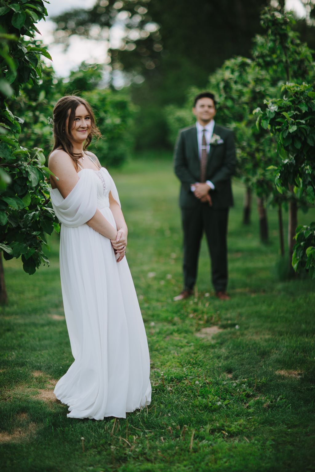 Pregnant bride in flowing white dress smiles at camera while groom stands blurred in background on lush green lawn