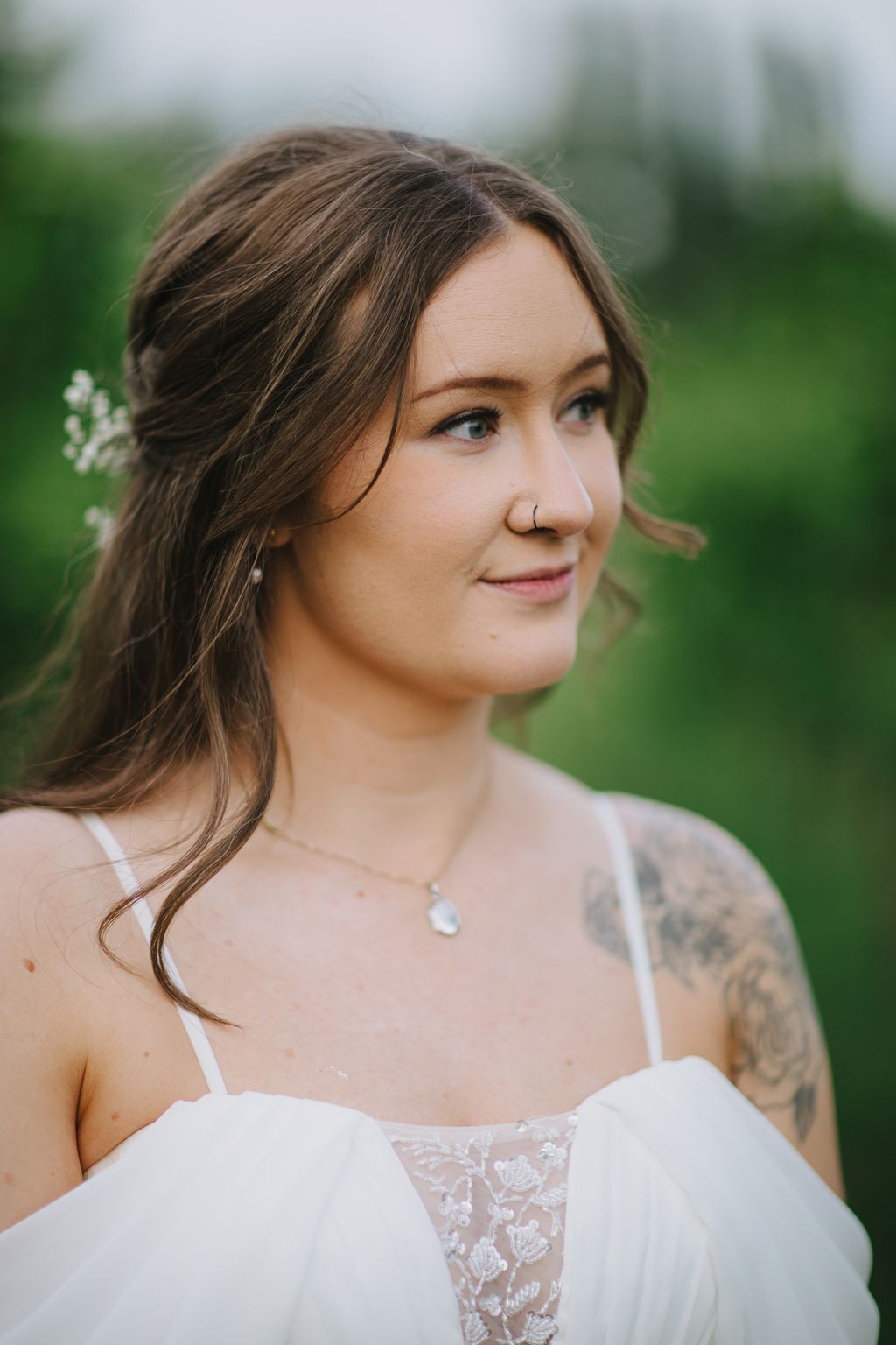 Bride in delicate lace dress with teardrop necklace gazes thoughtfully outdoors in soft, overcast light