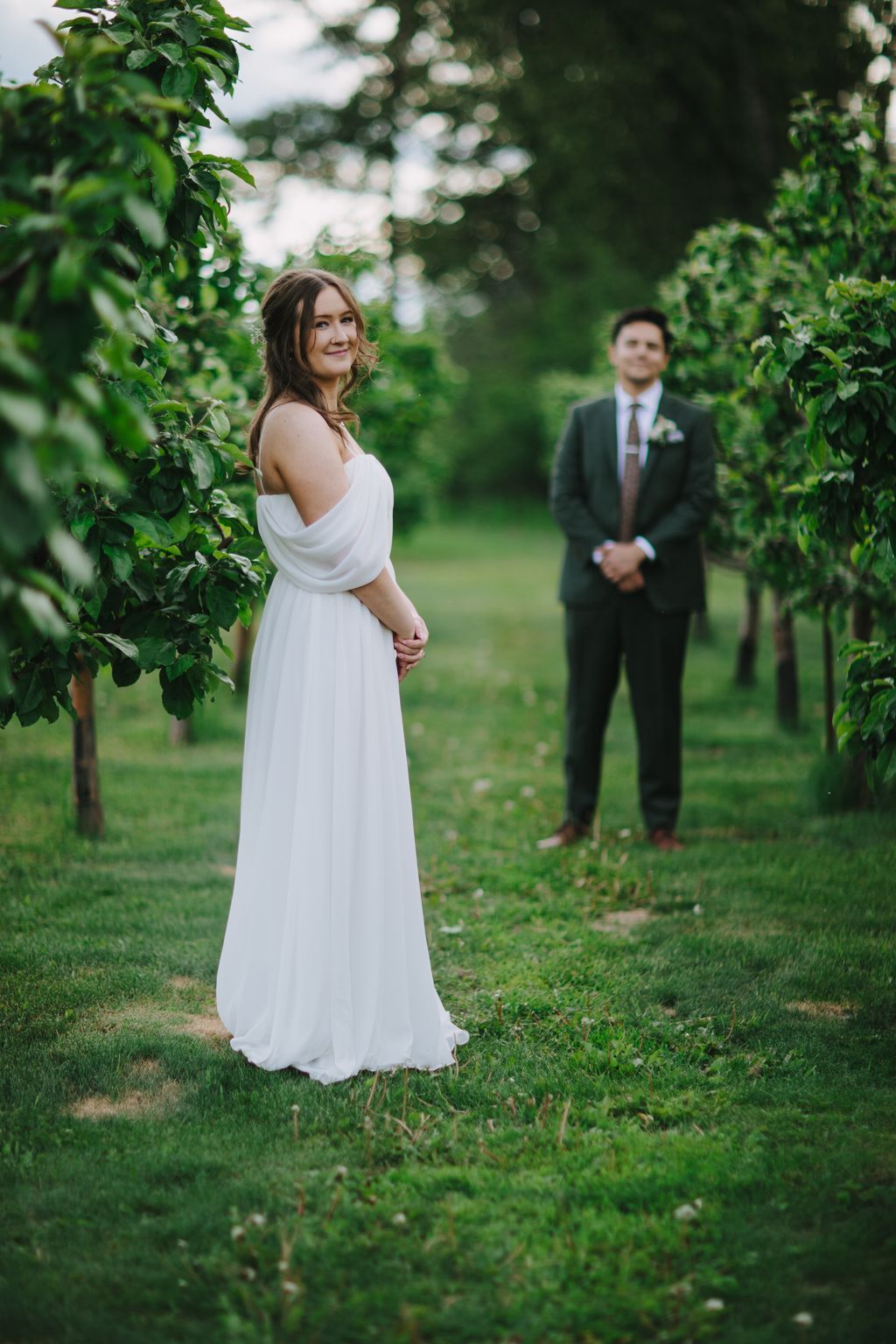 Bride in flowing white gown gazes over shoulder toward groom in dark suit across lush green lawn at golden hour