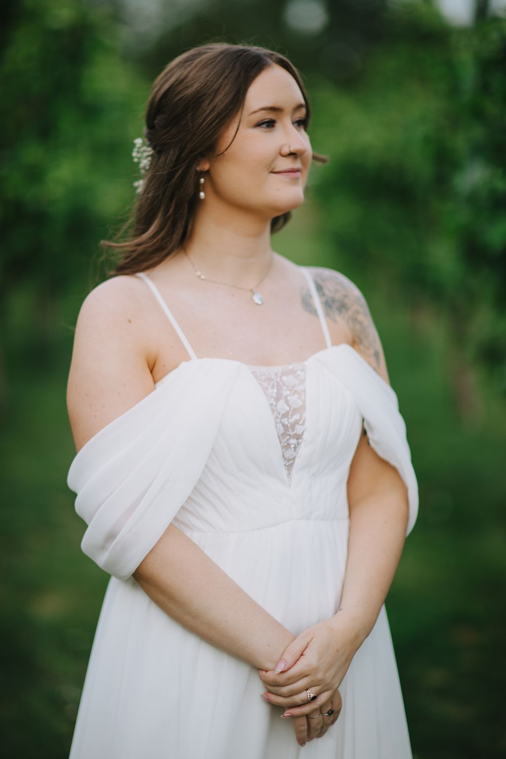 Pregnant bride in white flowing gown with lace detail gazes to the side, hand on belly, wearing pearl jewelry against soft green blurred background