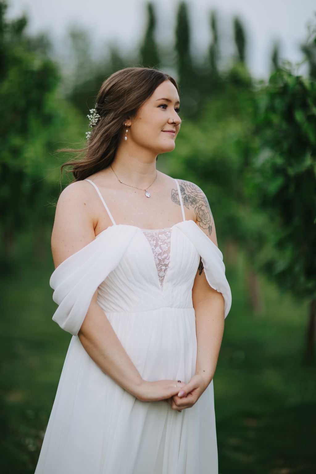 Pregnant bride in flowing white off-shoulder gown with baby''s breath in hair, standing in profile against blurred cypress trees