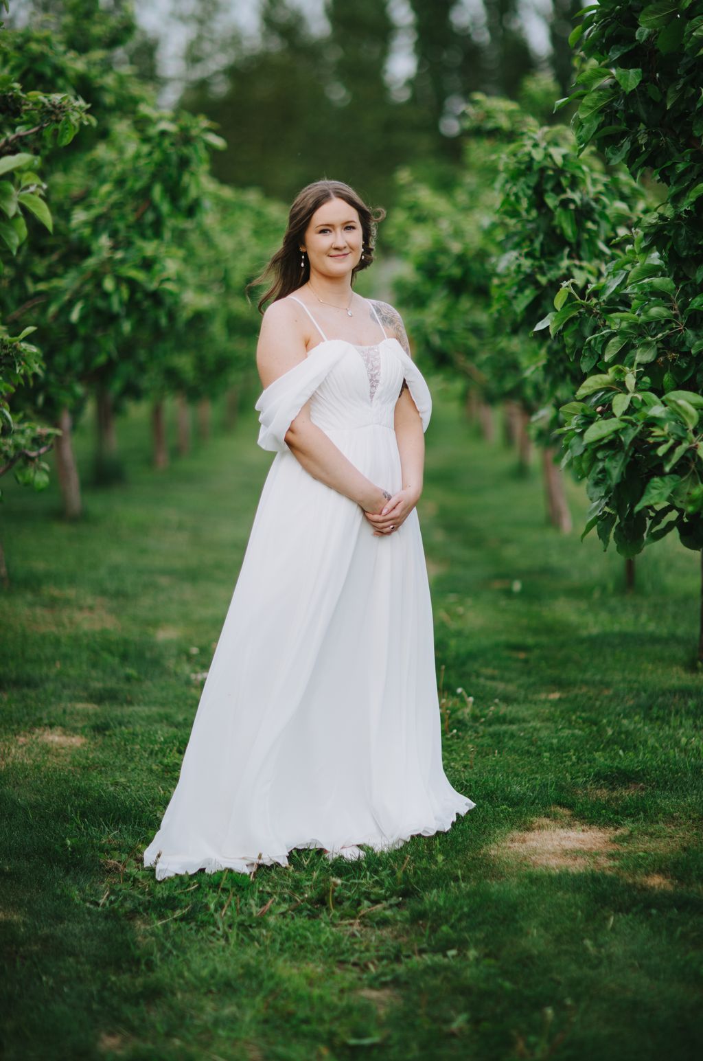 Pregnant woman in flowing white off-shoulder gown stands in lush green garden, hand on belly, soft natural light