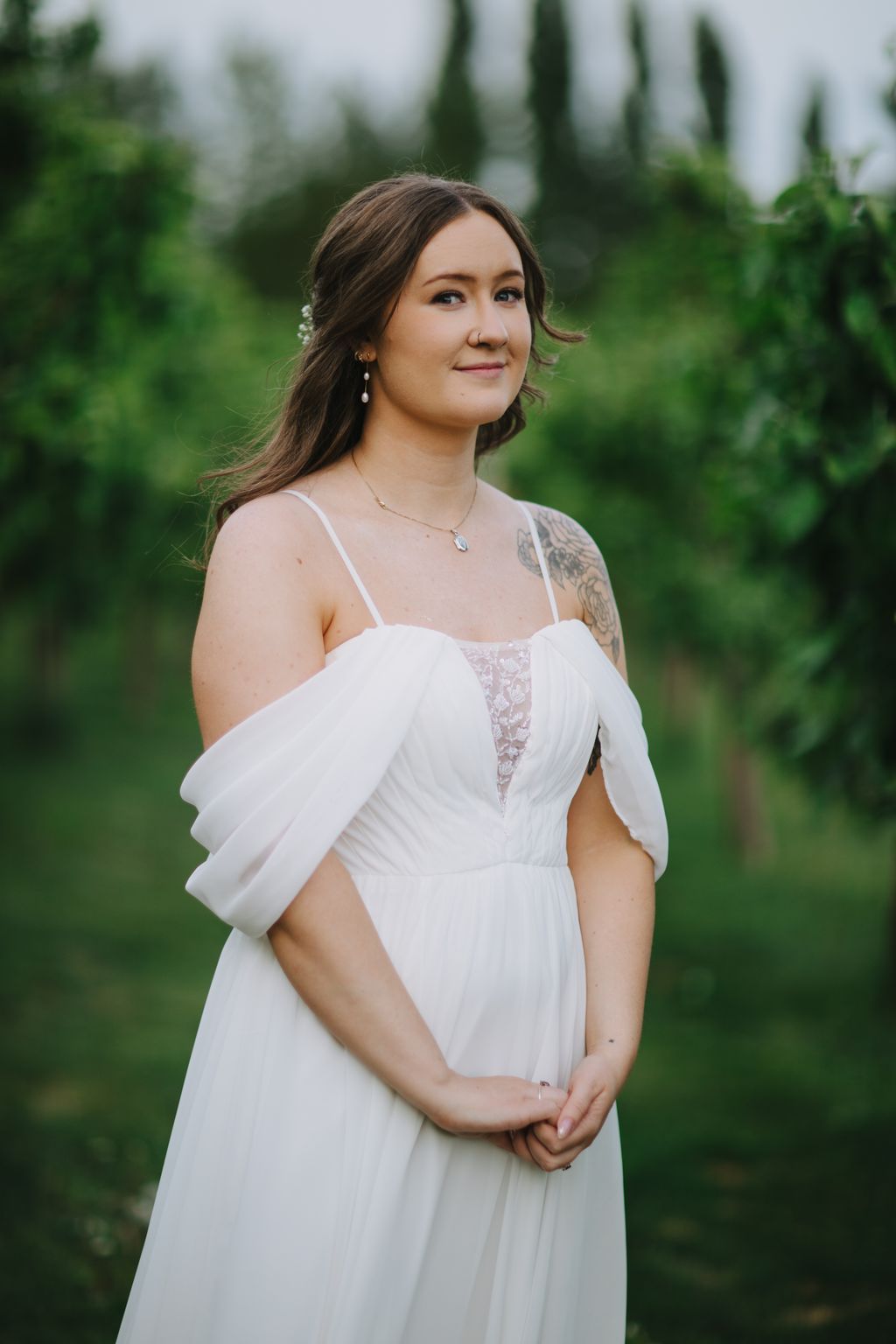 Bride in flowing white off-shoulder dress with lace detail gazes away, soft garden setting with cypress trees and overcast light