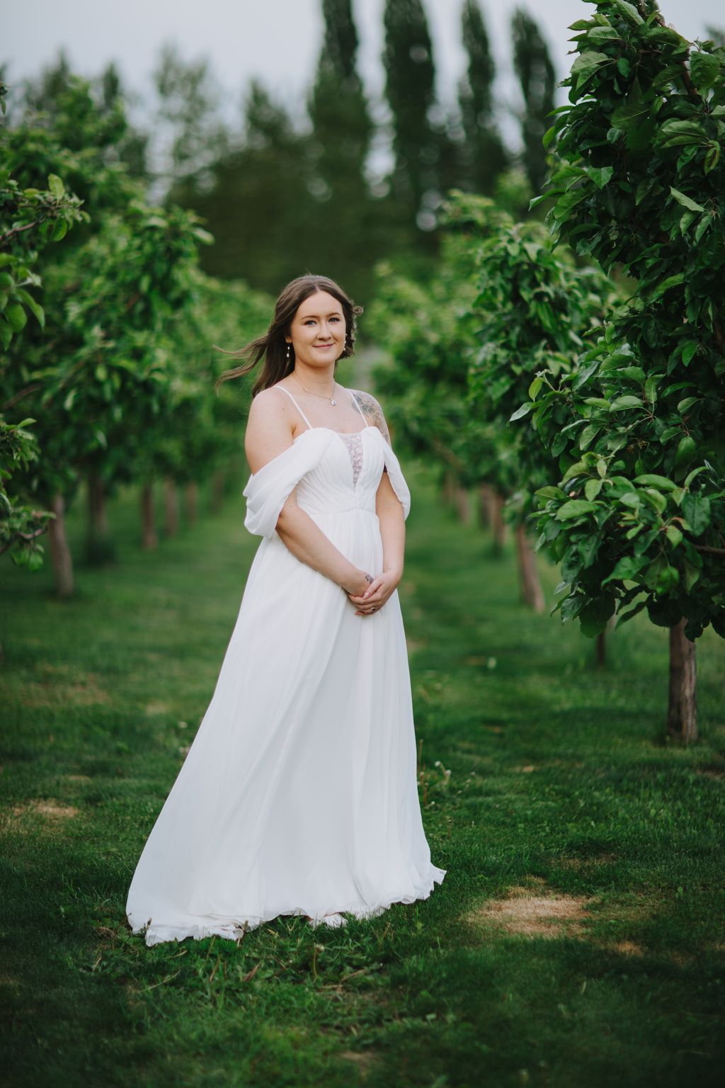 Pregnant woman in flowing white off-shoulder gown smiling in lush garden with tall evergreen trees