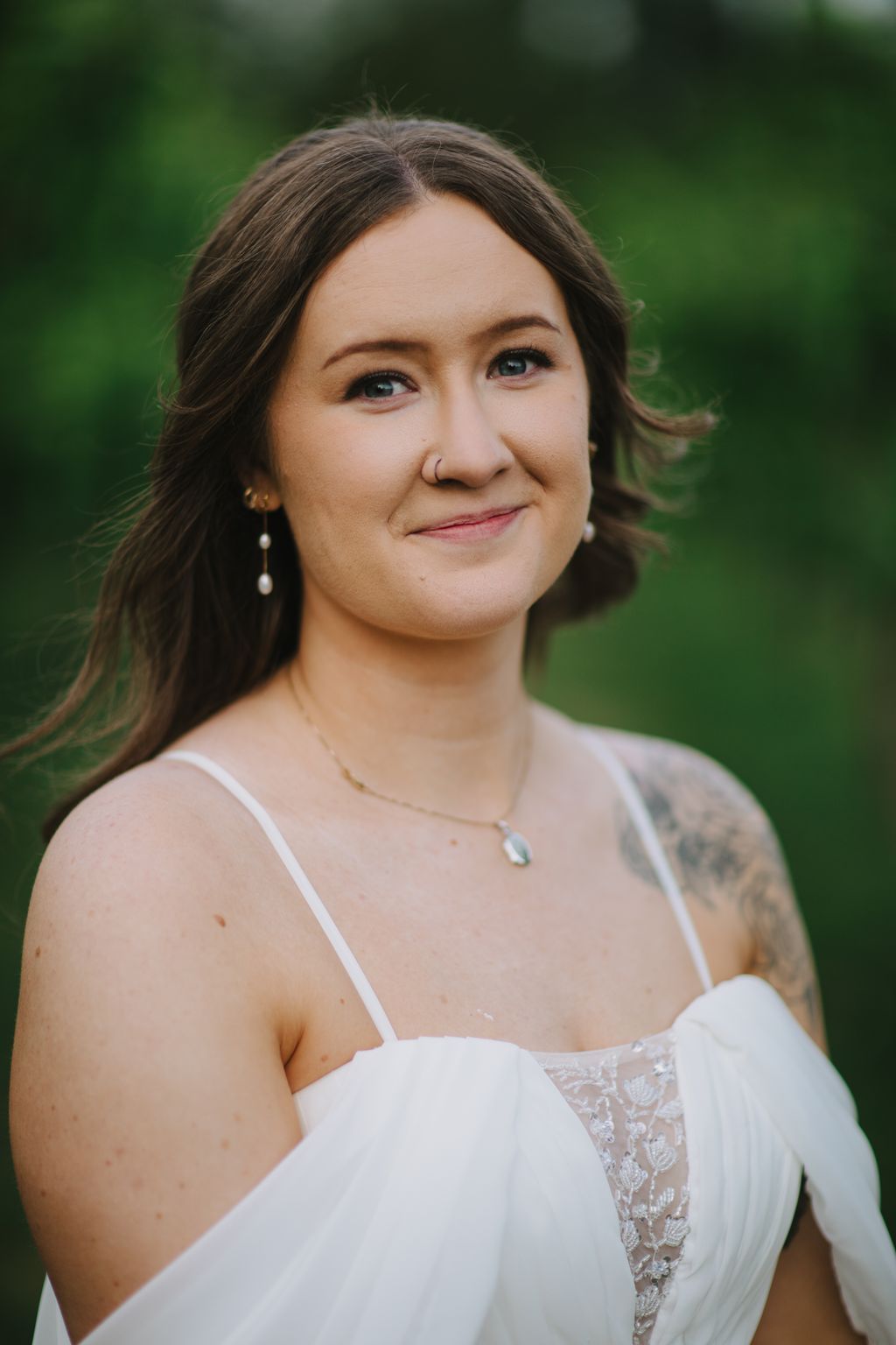 Bride in white lace gown with pearl earrings smiles softly against blurred green background