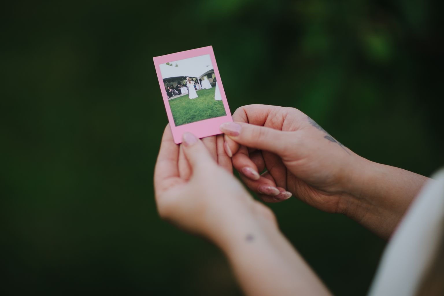 Hand holding pink Instax photo of bride and groom on grass against dark green background