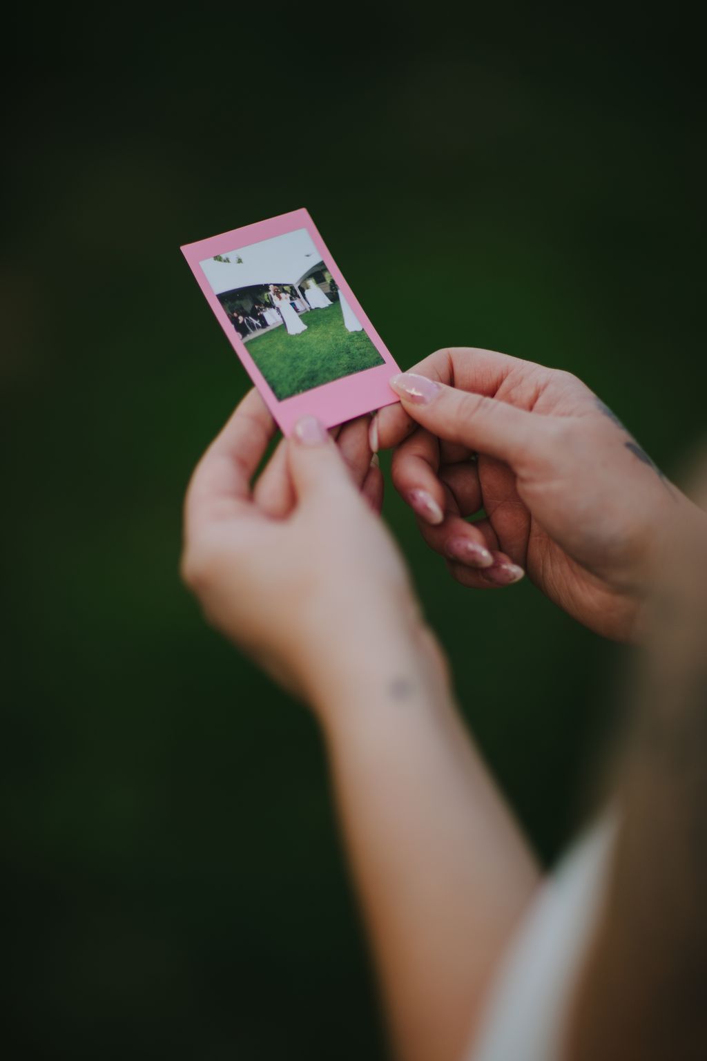 Hands holding a pink instant photo against a green background, showing a bride and groom on grass