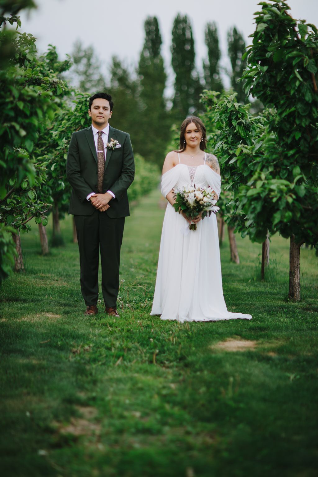Bride in flowing white off-shoulder gown and groom in dark suit stand together in lush vineyard with tall cypress trees under overcast sky