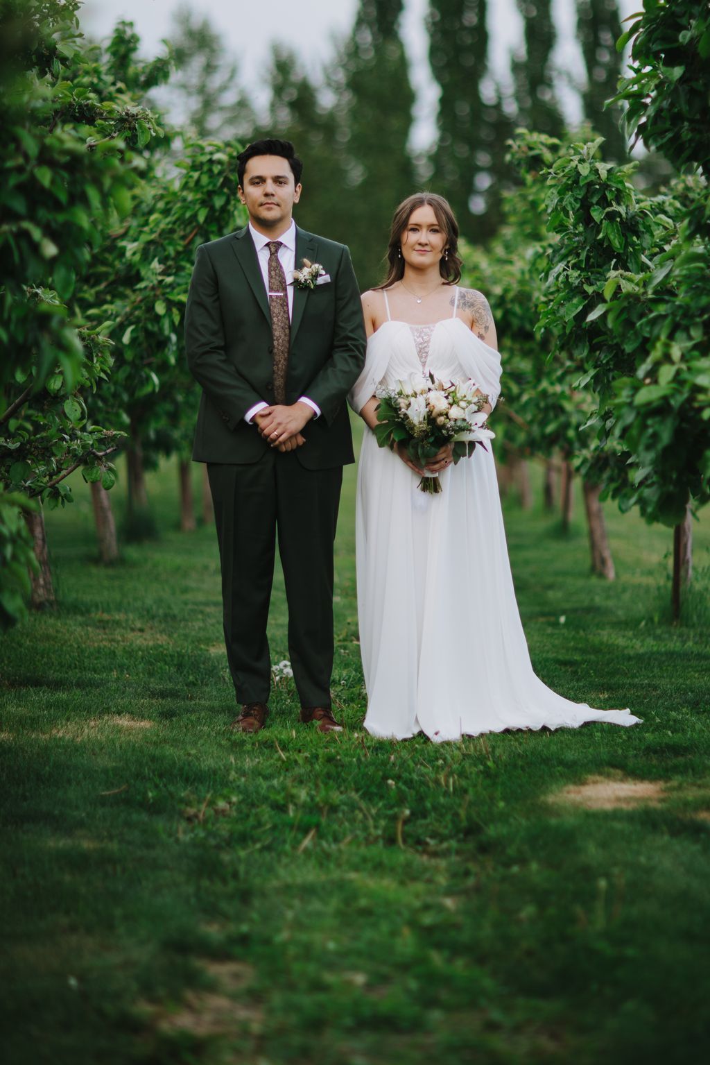 Bride in flowing white dress holding organic bouquet stands beside groom in dark suit against evergreen trees and grass