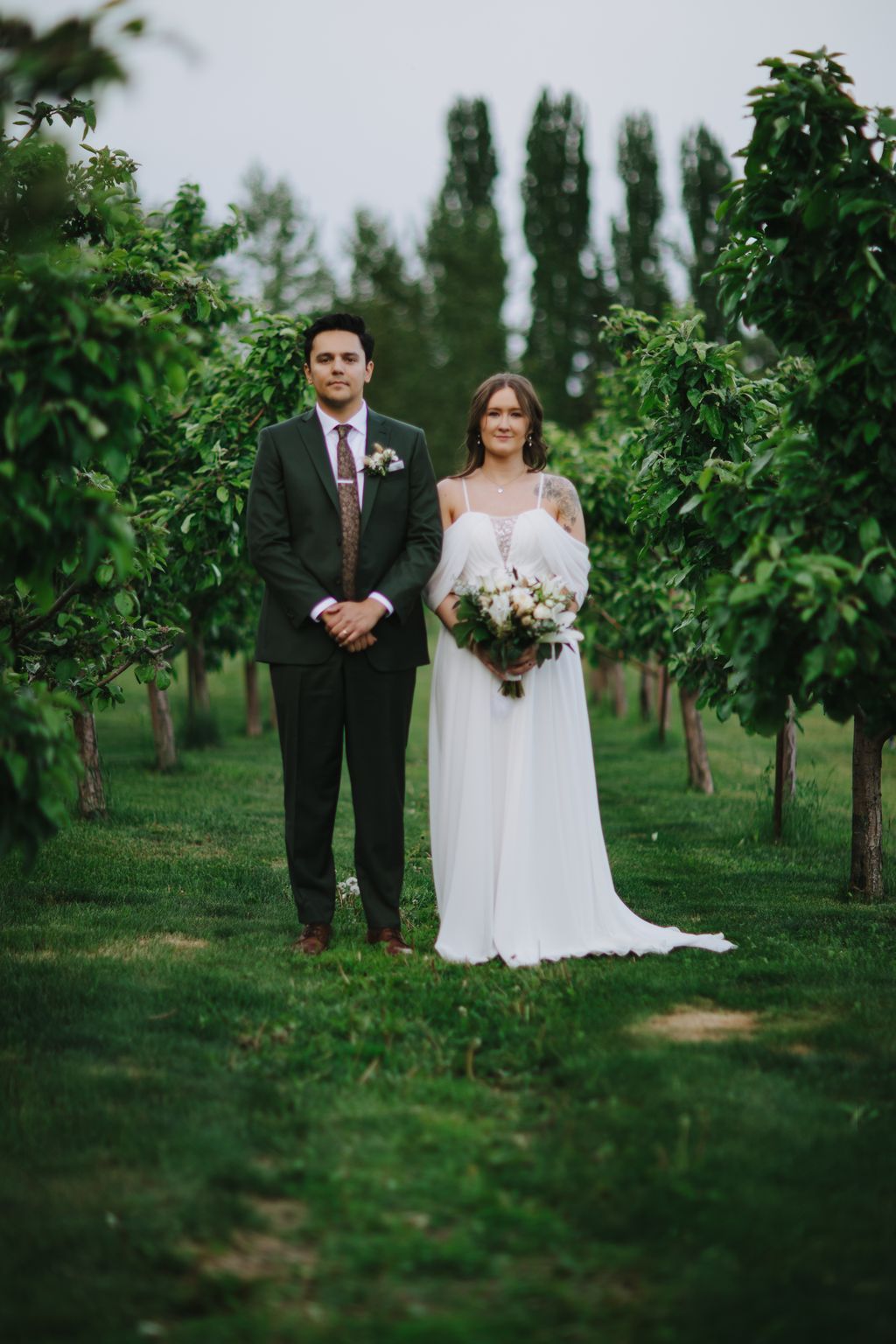 Bride in white off-shoulder dress and groom in dark suit stand together in lush garden with tall cypress trees and ivy-lined path